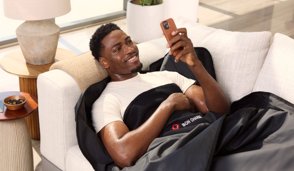 man laying on sofa in a sauna blanket while using his phone