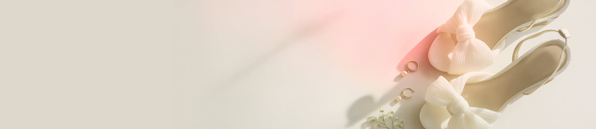 Pair of white high-heeled shoes with decorative elements on a light pink background, next to a set of wedding rings.